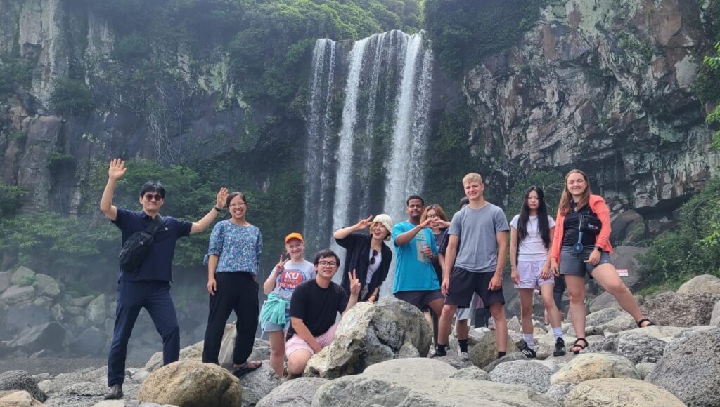 From left: Joo Yong Kim, Stacy Nam, Erica Hunsberger, Andrew Hoang, Sunju Moon, Biz Bomberger, Eunjin Kim, Tommy Nguyen (hidden), Ian Hunsberger, Hannah Kim, and Joanna Spiker visited a waterfall near Gangjeong on Jeju Island in July 2024. Photo by Kaia Vereide.