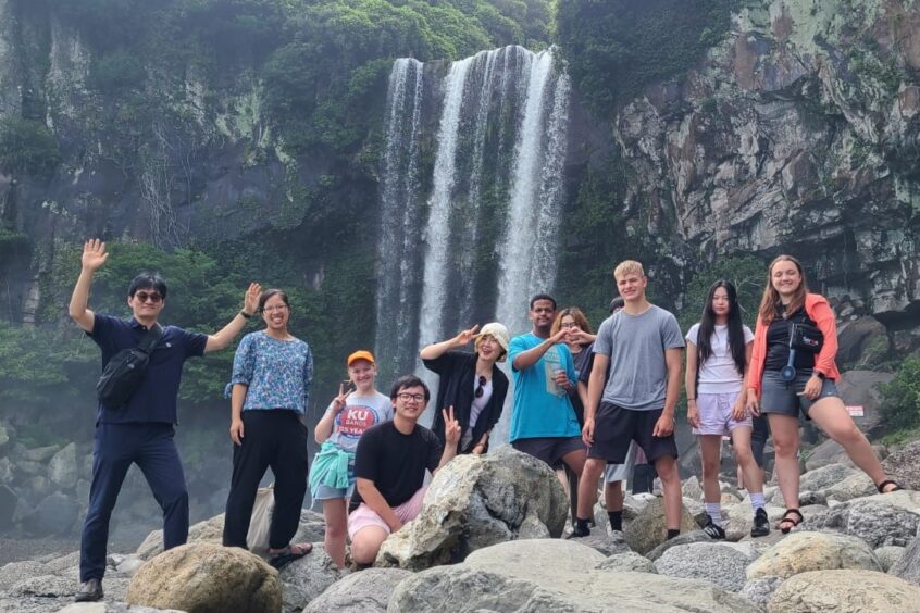 From left: Joo Yong Kim, Stacy Nam, Erica Hunsberger, Andrew Hoang, Sunju Moon, Biz Bomberger, Eunjin Kim, Tommy Nguyen (hidden), Ian Hunsberger, Hannah Kim, and Joanna Spiker visited a waterfall near Gangjeong on Jeju Island in July 2024. Photo by Kaia Vereide.